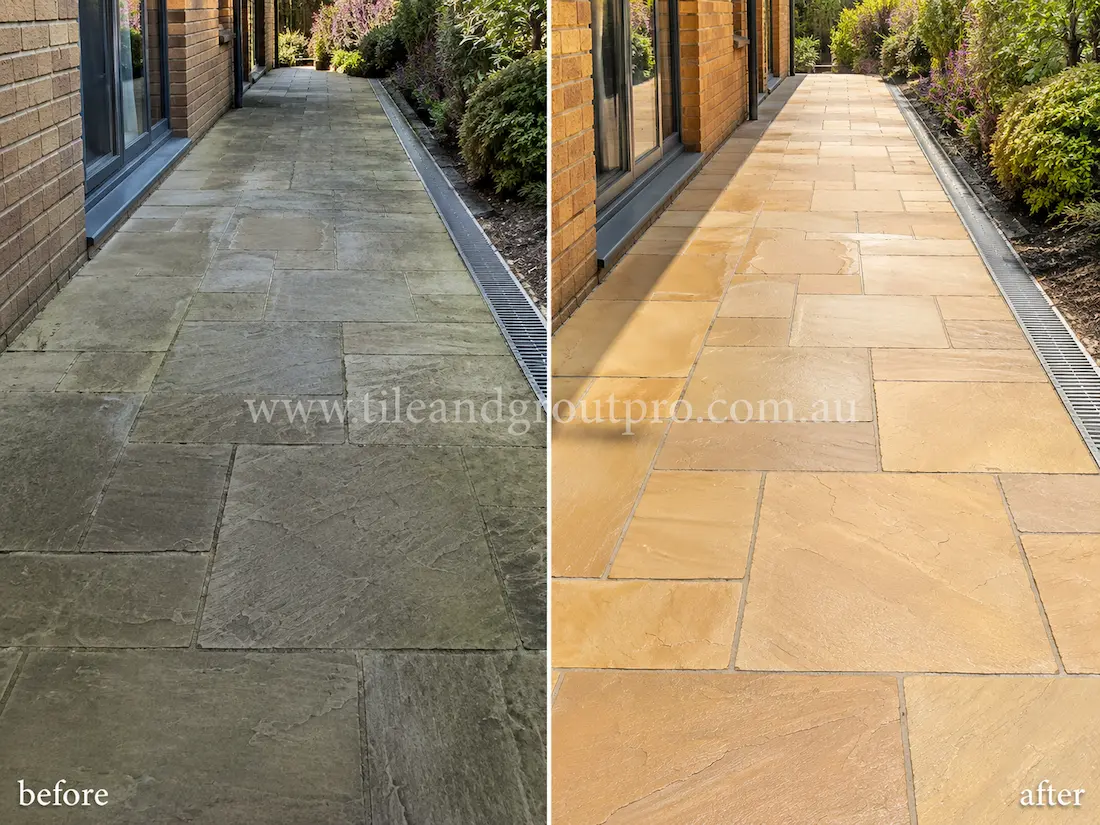 Before tile renewal, outdoor sandstone pathway tiles along a residential side yard walkway appeared weathered with dark staining, surface grime, and uneven natural stone colouring beside landscaped garden beds.